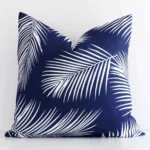 Navy palm-designed cushion cover sits against a white wall. It is constructed from a superior looking outdoor material and has square dimensions.