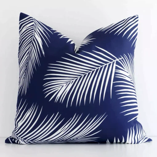 Navy palm-designed cushion cover sits against a white wall. It is constructed from a superior looking outdoor material and has square dimensions.