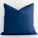 A blue, solid-coloured cushion cover sits against a white wall. It is constructed from a superior looking outdoor material and has square shape.