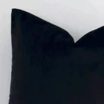 Enlarged shot of the corner of this square solid-coloured cushion cover in black colour is shown against a brick wall. The image shows the quality and craftsmanship of the velvet material.