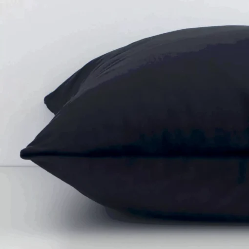 A black solid-coloured cushion arranged sideways in front of a wall. The square shape and velvet fabric are shown and the seams are clearly visible.