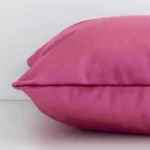 A solid-coloured blush cushion cover is laid flat. This angle shows the side of the velvet fabric and its square shape.