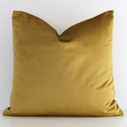 A white wall that has a solid-coloured gold mustard cushion cover positioned in front of it. It has an exquisite velvet material and a lovely square shape.