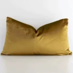 A rectangle solid-coloured cushion in a delightful gold mustard tone rests against a white wall. The velvet material appears to be of exceptional quality.